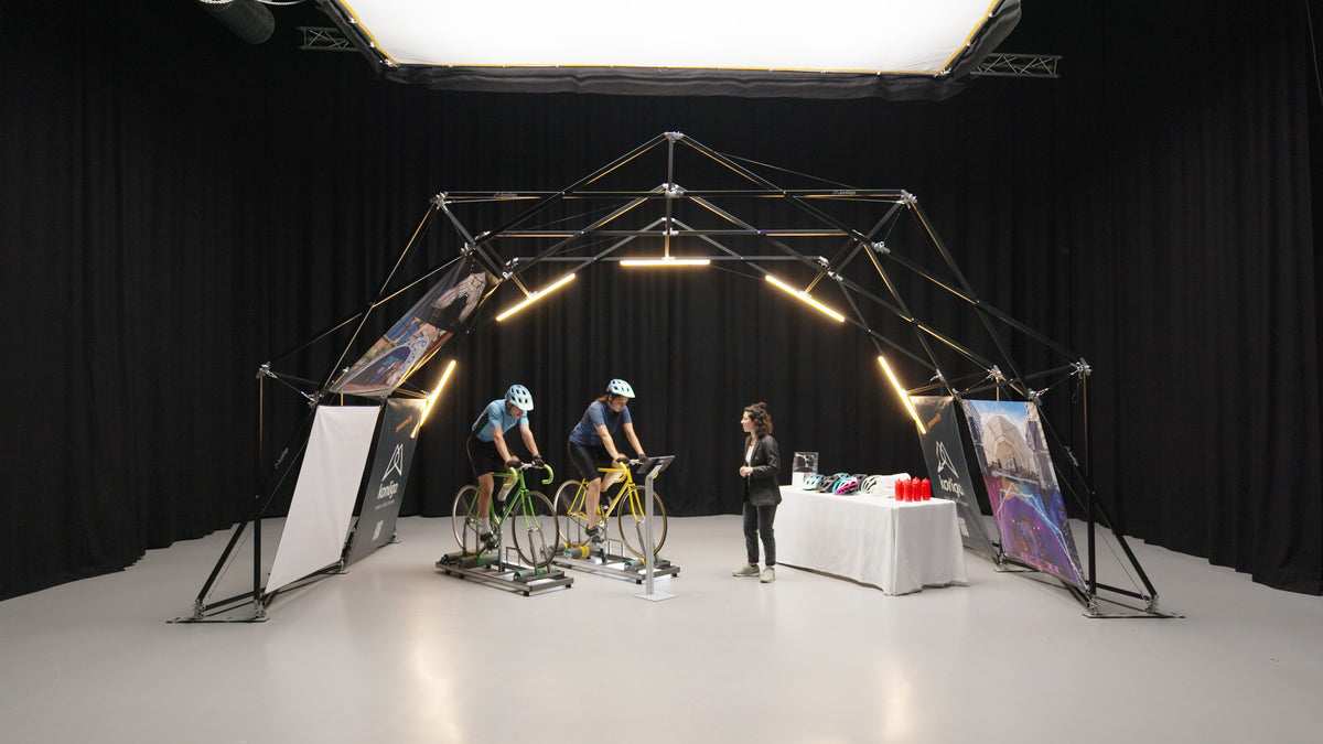 Configuración interior de la estructura para eventos de Konligo con dos ciclistas en bicicletas estáticas y una mesa de exhibición con cascos y botellas de agua. Ahora disponible en MTN Shop EE. UU.
